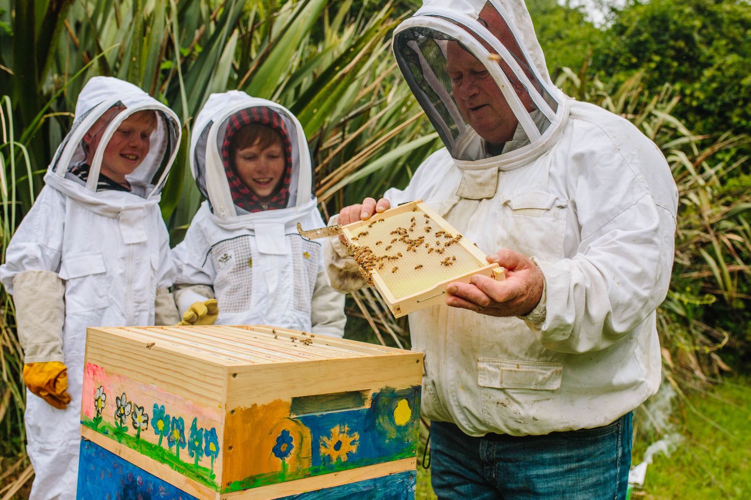 Beekeepers in protective suits examining honeycomb frame from wooden hive box.