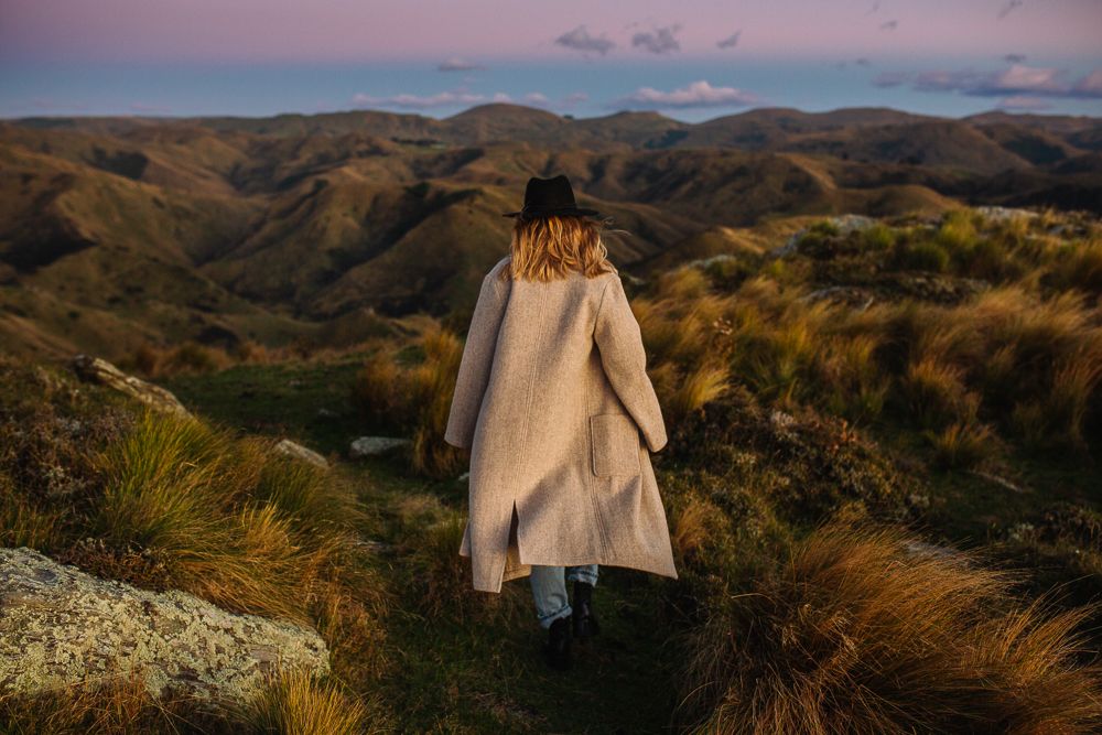 Person in beige coat overlooking mountainous landscape at sunset.