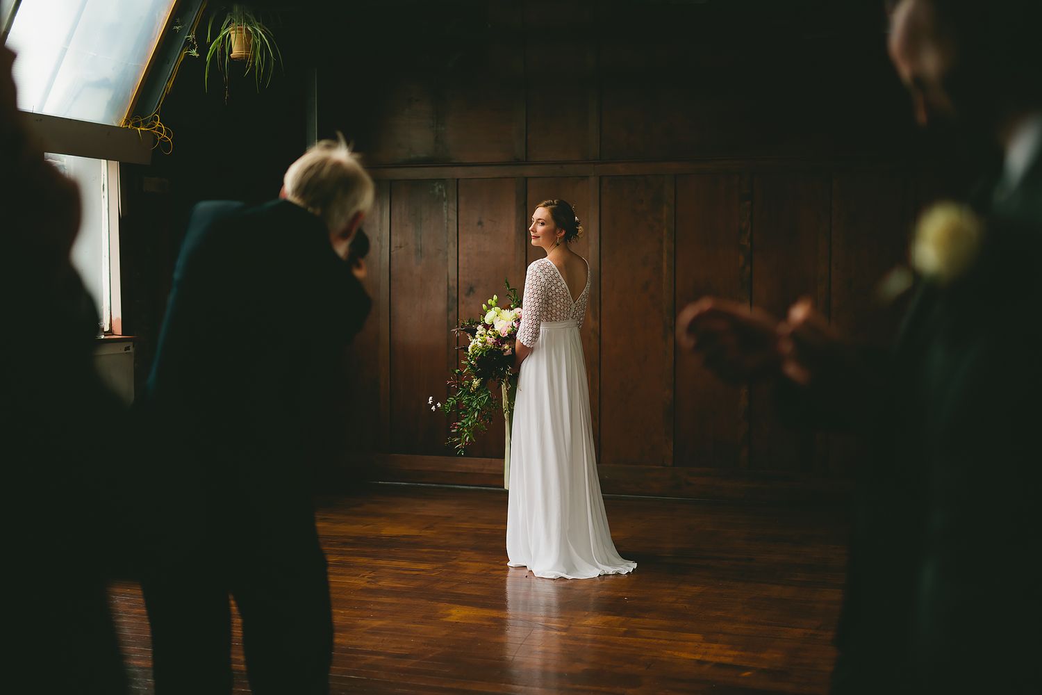 A bride in a flowing white wedding gown stands in a grand wooden-paneled room during a wedding ceremony.