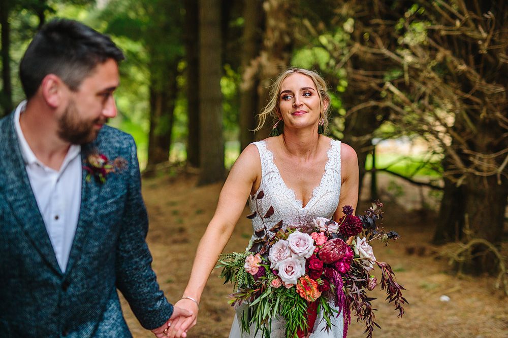 Wedding couple walking through lush green forest holding vibrant burgundy floral bouquet.