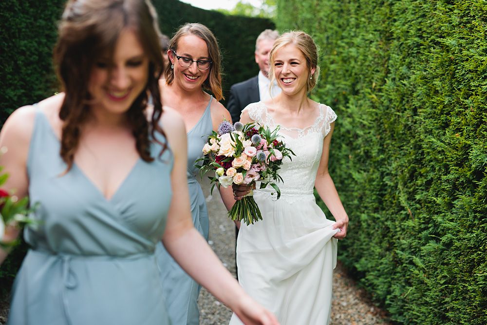 Wedding party members in light blue dresses walk through a lush garden pathway.