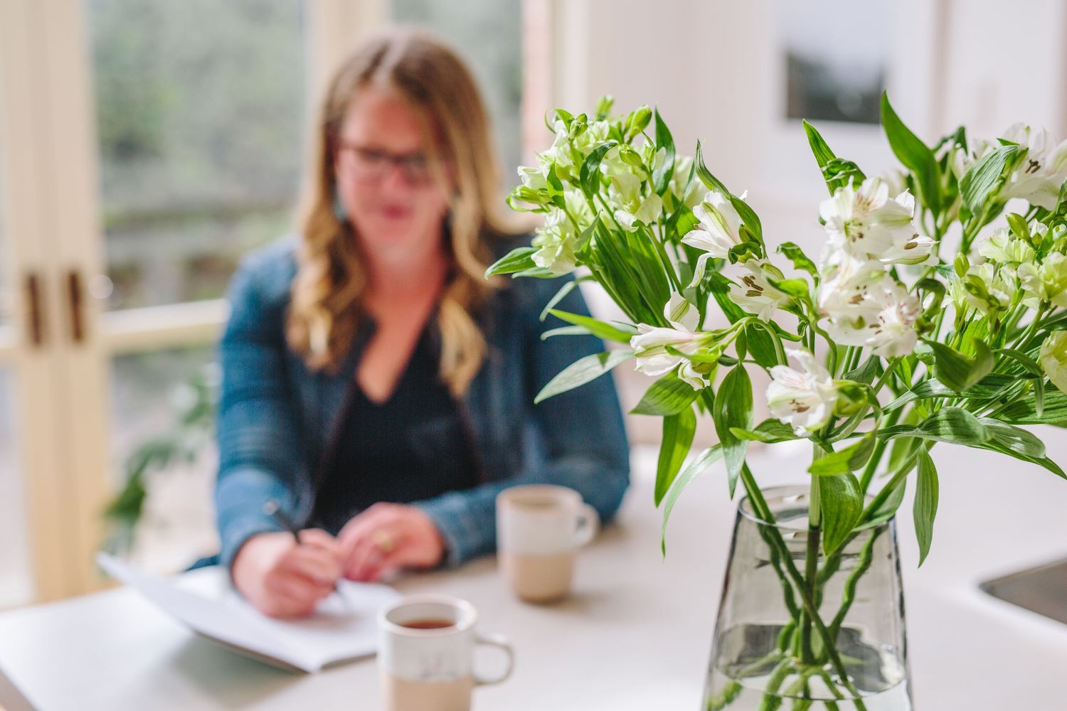 People gathered around a table with coffee cups and green plant decorations in a bright, modern workspace setting.
