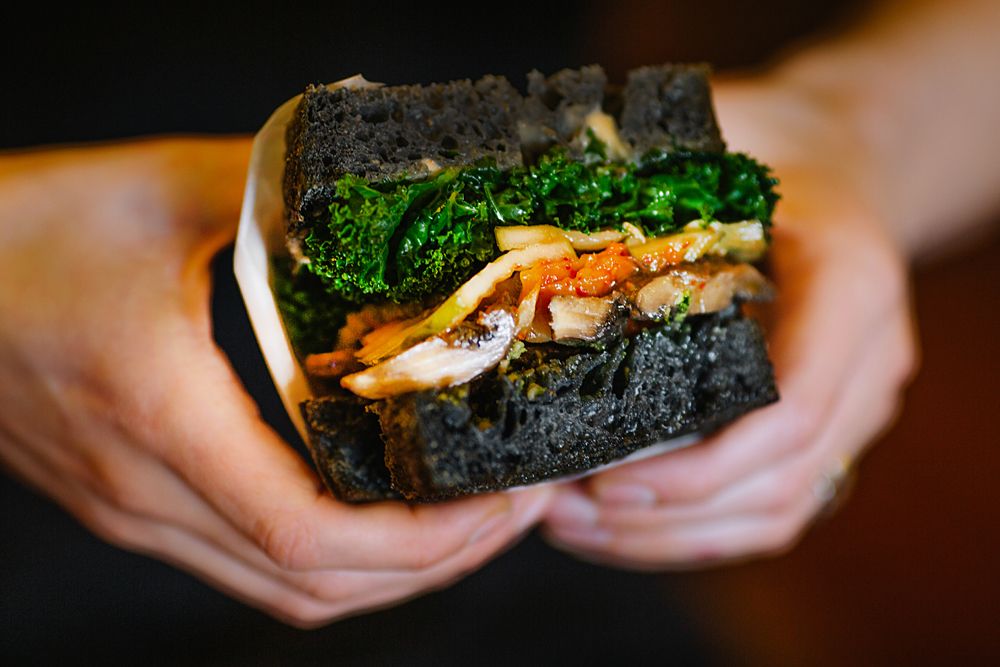 Black charcoal sandwich with green vegetables and meat filling held in hands.