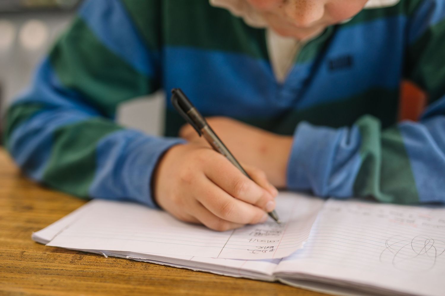 Close-up view of hands writing with a pencil on white paper at a wooden desk surface.