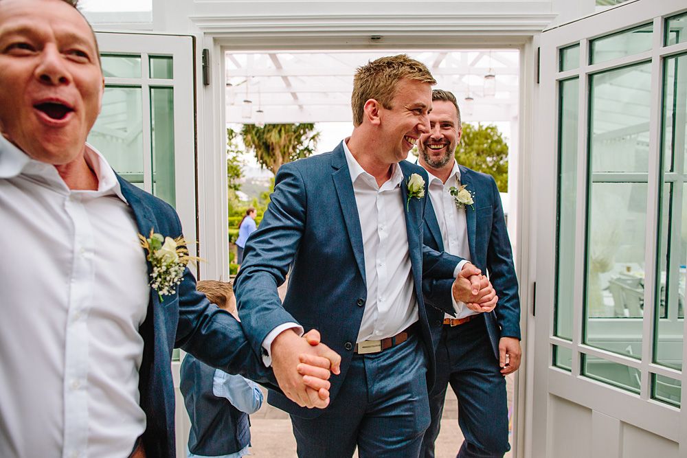 Wedding party shares laughter and joy in bright sunlit conservatory.