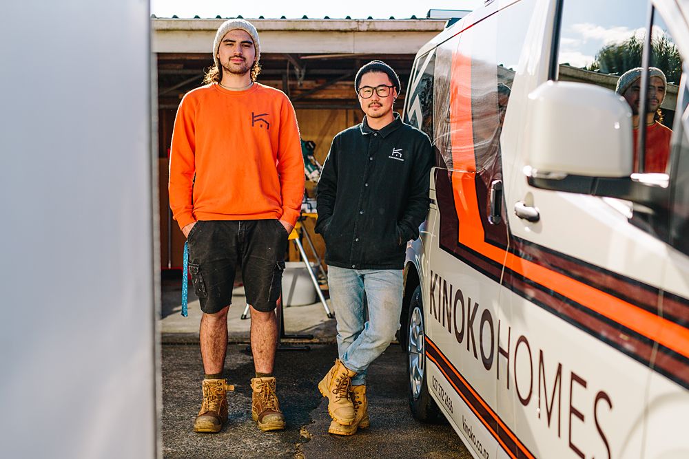 Two workers in orange and black workwear stand in front of a company vehicle.
