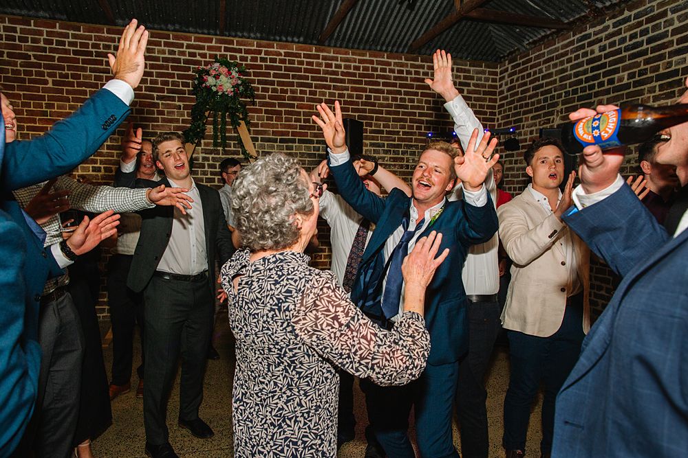 Wedding party dancing with hands in the air against rustic brick wall backdrop.