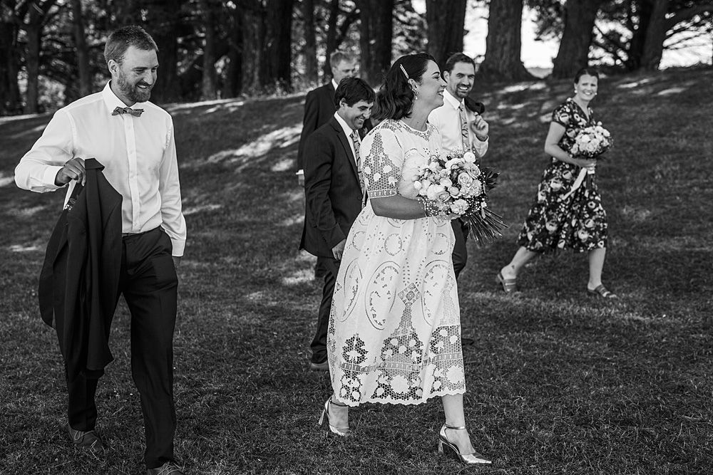 Wedding party members walking casually through a sunlit garden setting.