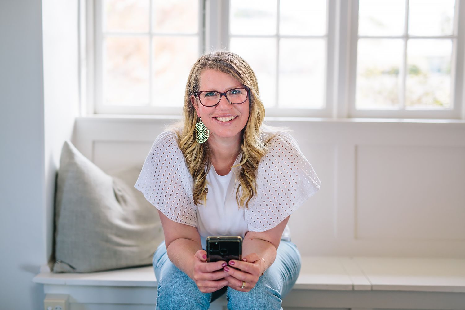 Person in glasses relaxes on white couch while using smartphone in bright, sunlit living room.