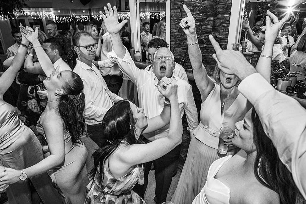 Wedding guests dance and celebrate with raised hands on a lively dance floor at an indoor reception.