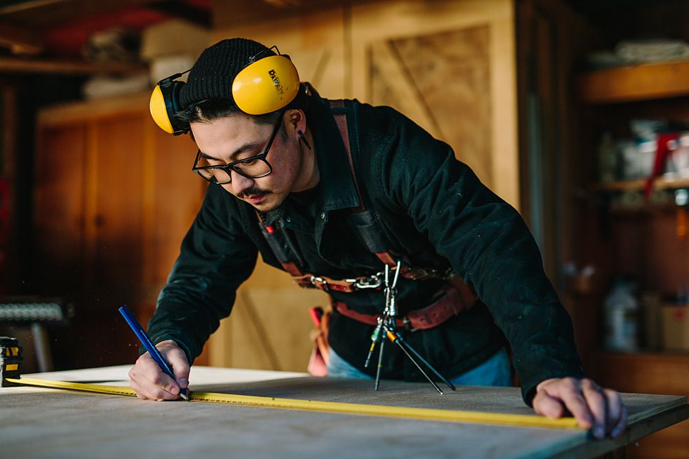 Person wearing protective ear muffs and glasses measures and marks on a workbench.