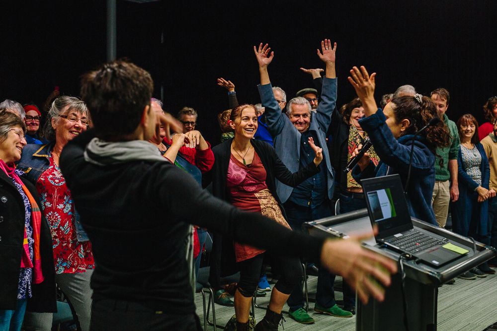 Enthusiastic crowd raises hands in celebration during an indoor event with a performer on stage.