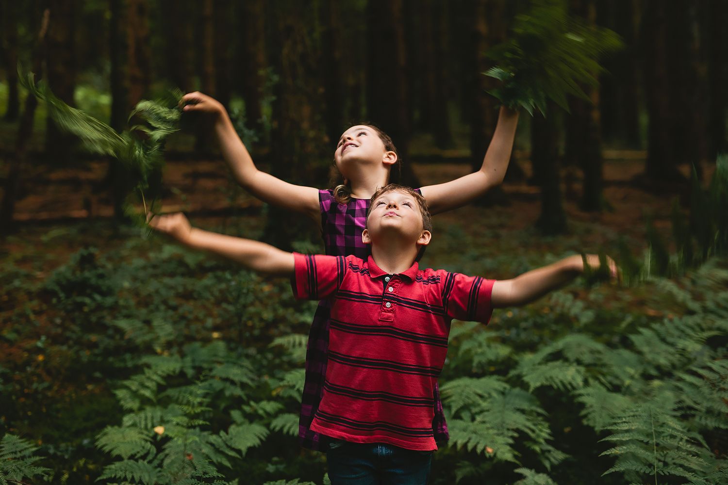Two children with arms raised playing in a lush forest with ferns.