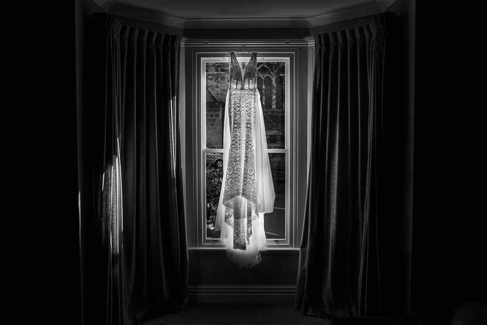 Wedding dress hangs elegantly in window light creating dramatic silhouette.