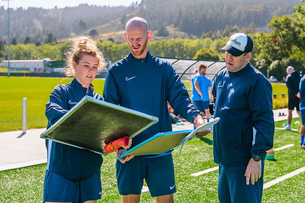 Team members in blue athletic gear reviewing plans on outdoor sports field.