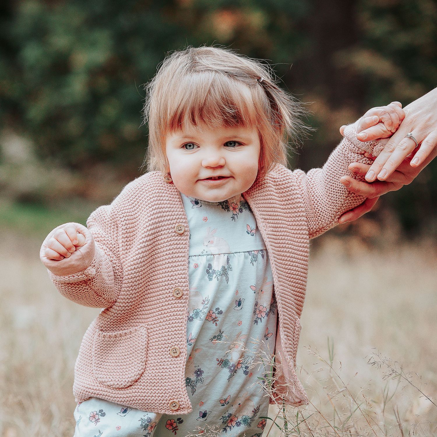 Baby girl taking her first steps through a garden setting in a nursery - she his holding on to a preschool teachers hand.