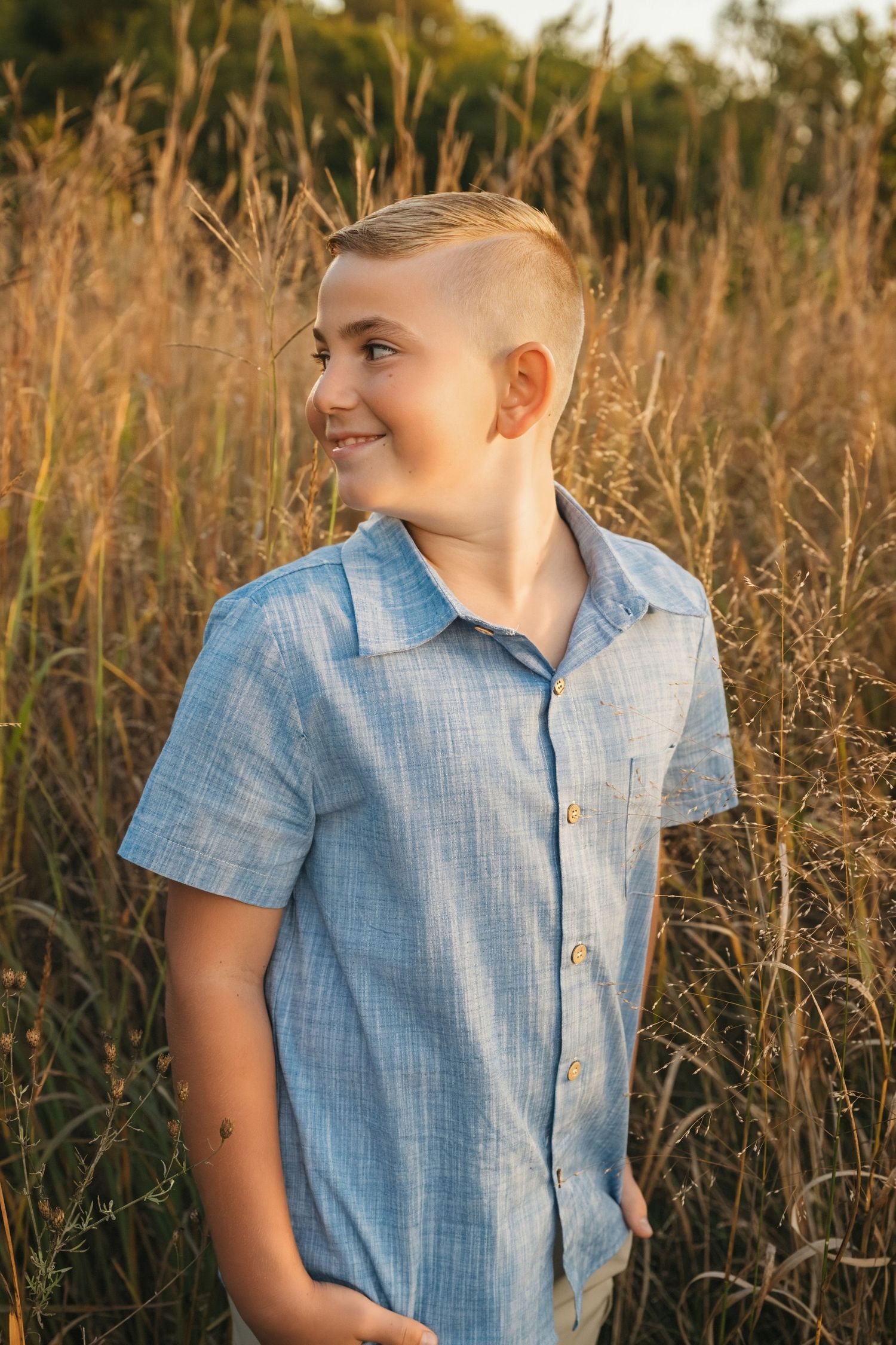 Smiling blonde boy in blue button-up shirt standing in tall golden grass field during sunset.