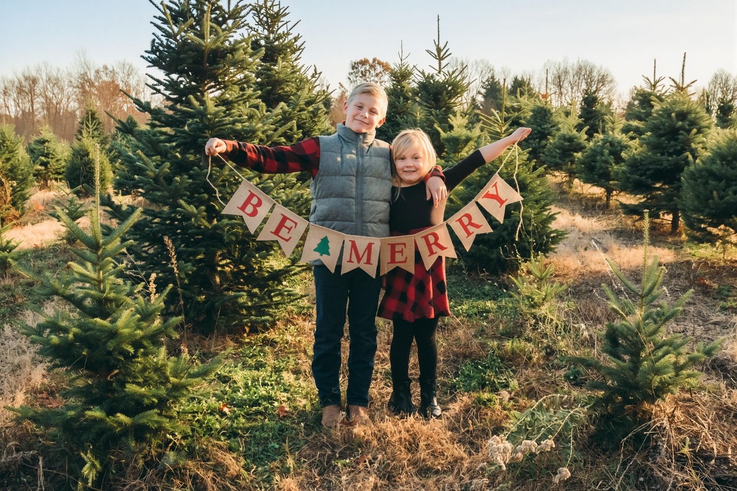 Two children holding a BE MERRY burlap banner at a Christmas tree farm surrounded by evergreen trees.