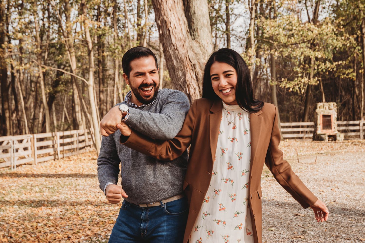 A cheerful couple laughing and posing outdoors in a wooded autumn setting, woman in floral dress and brown coat.