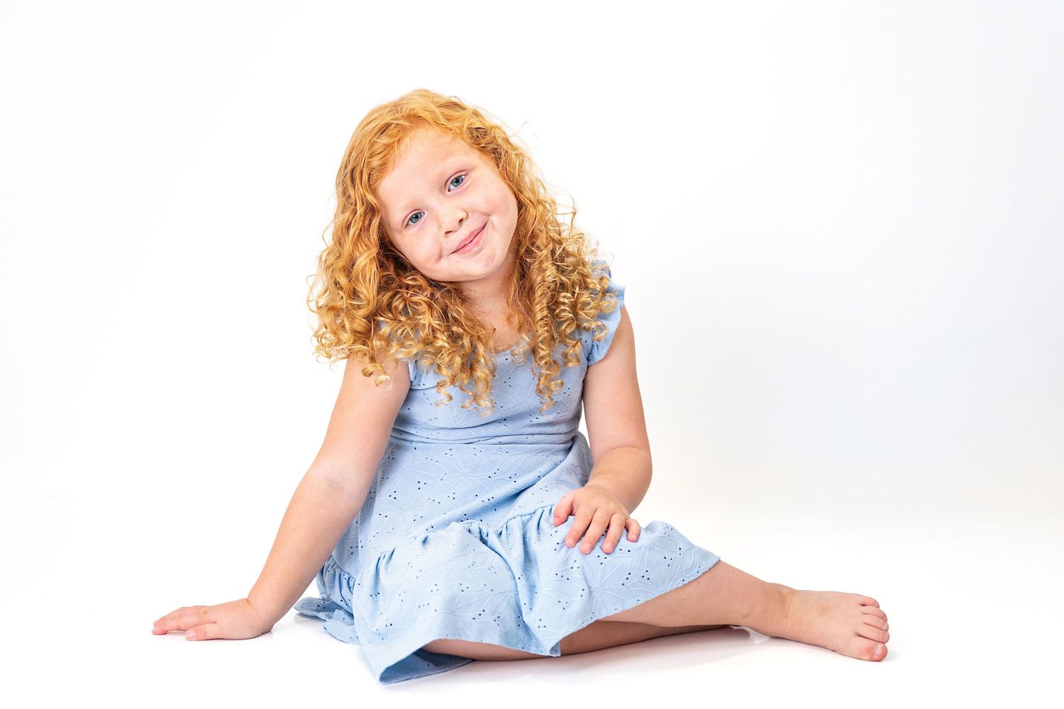 Adorable young girl with curly red hair in a blue dress sitting and smiling on white background.