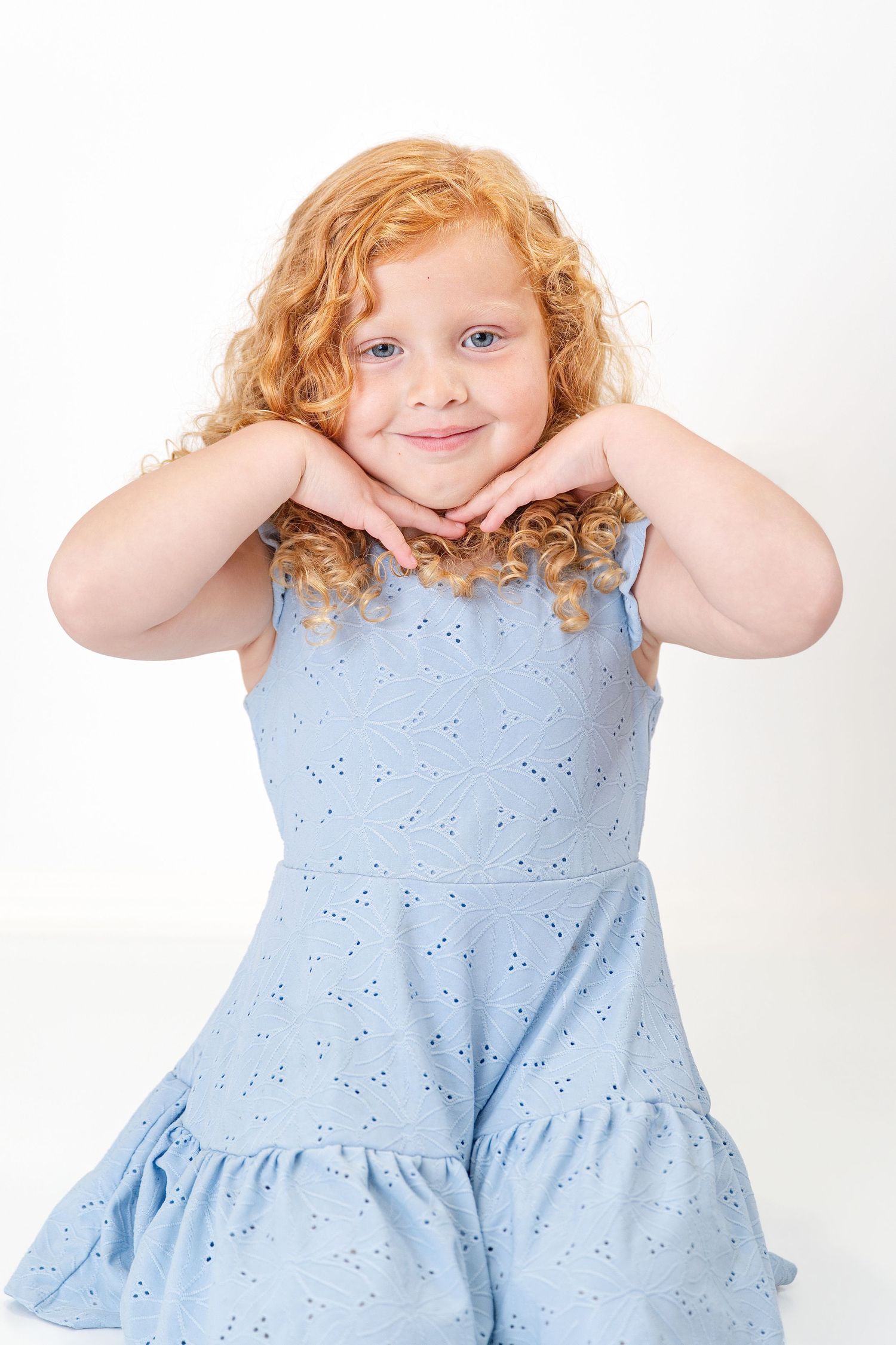 Adorable young girl with curly red hair wearing a blue dress, posing with hands under chin against white background.