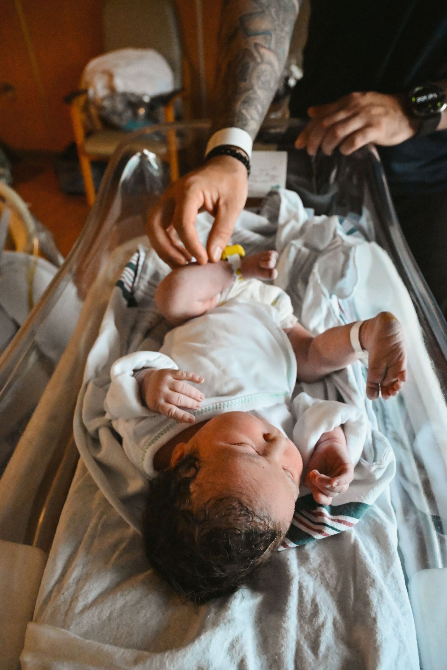 Newborn baby lying in hospital bassinet while parent touches infant gently with tattooed hand.