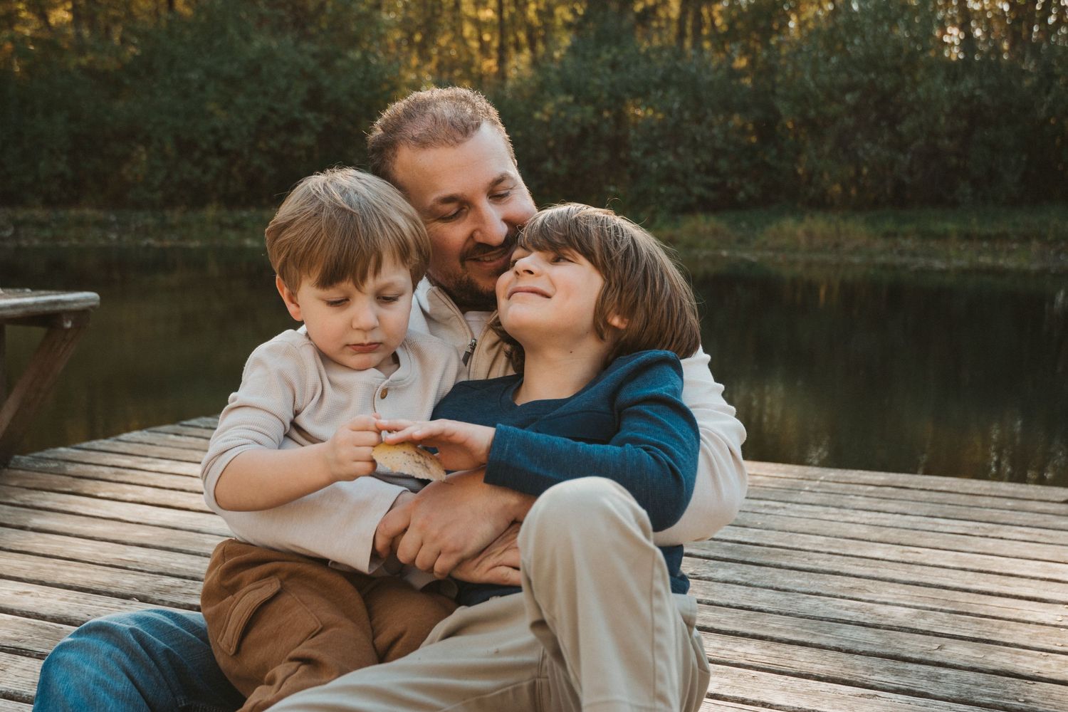 Candid outdoor family photo of dad with his two sons sitting on a dock, captured during an emotional Macomb, Michigan session