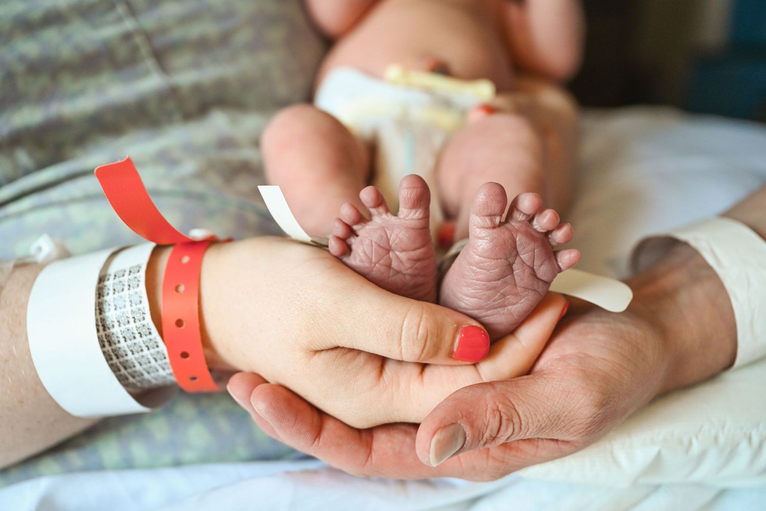 Parents holding tiny newborn baby feet in hospital, with colorful ID wristbands visible on their wrists.