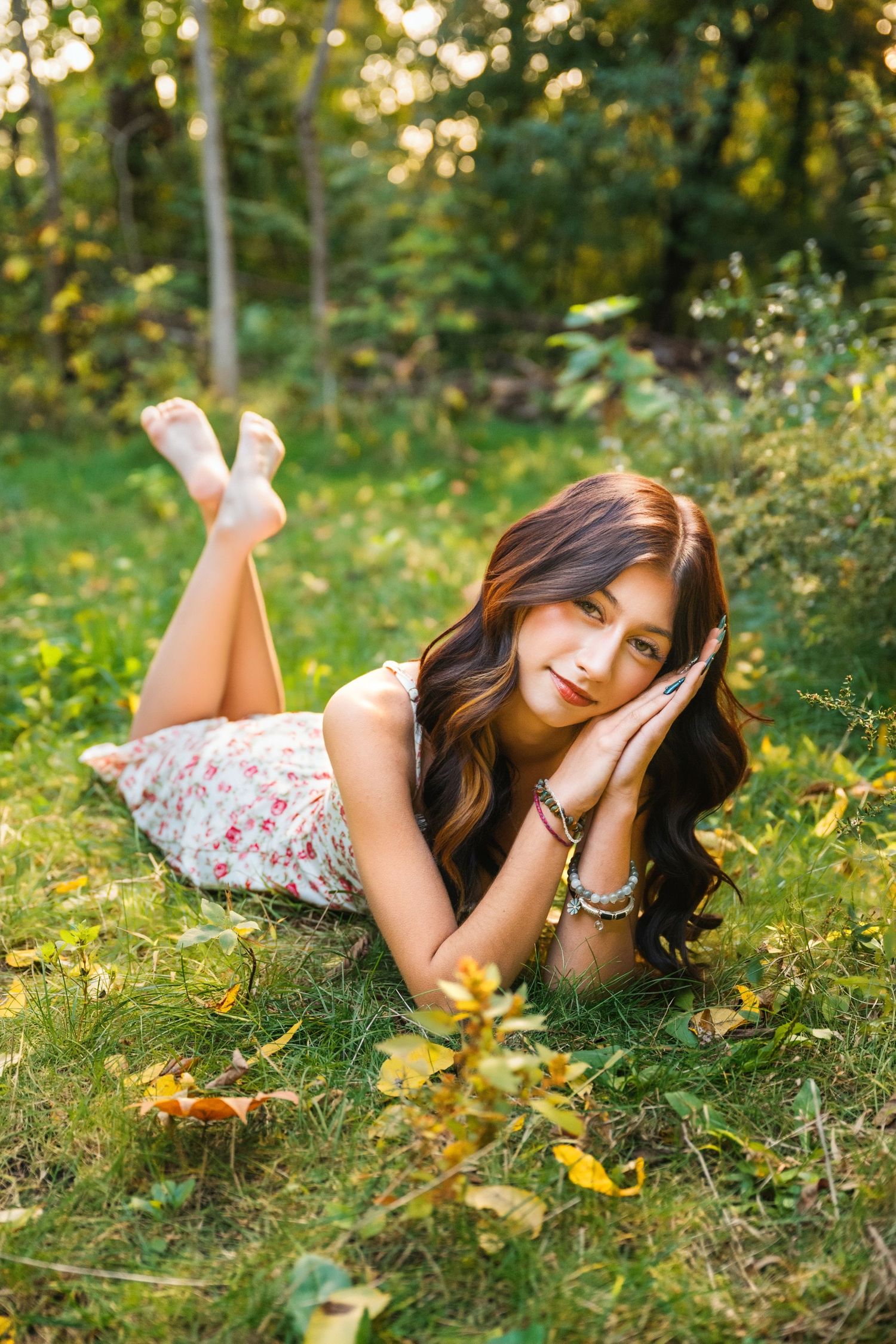 Young woman with long dark hair lying on green grass in a floral dress, relaxing outdoors in a sunny forest setting.