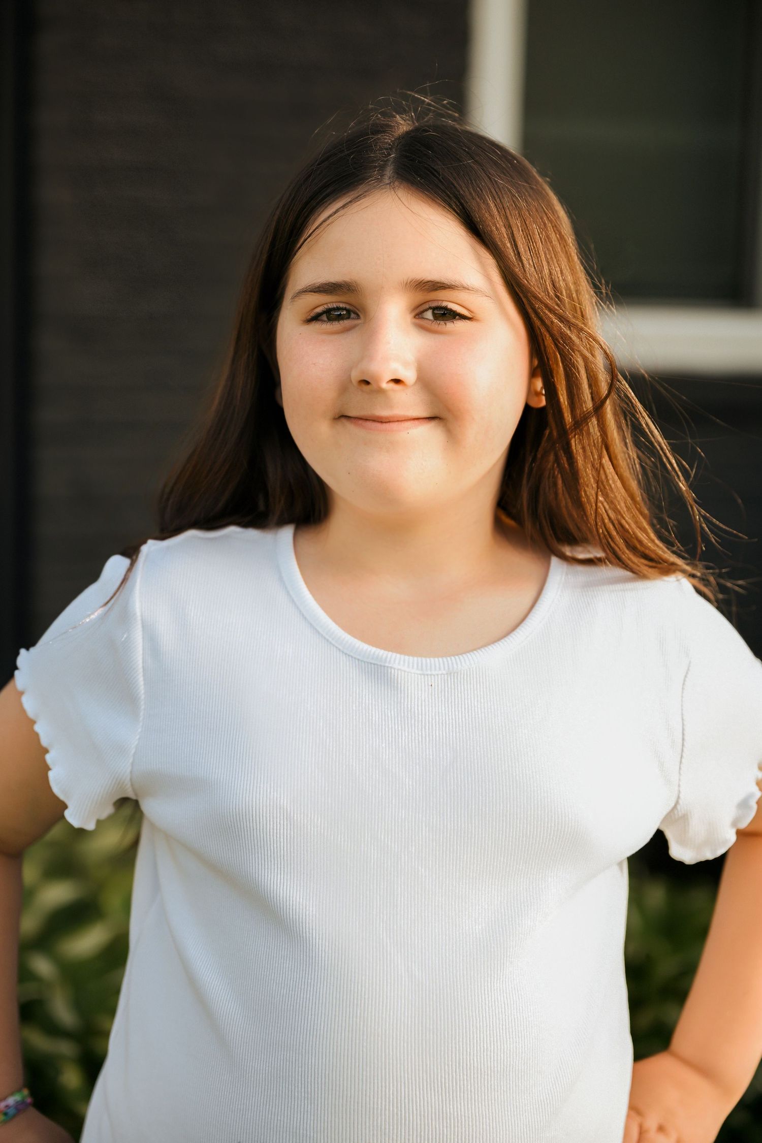 Young girl with long brown hair smiles confidently outdoors wearing a white short-sleeve t-shirt.