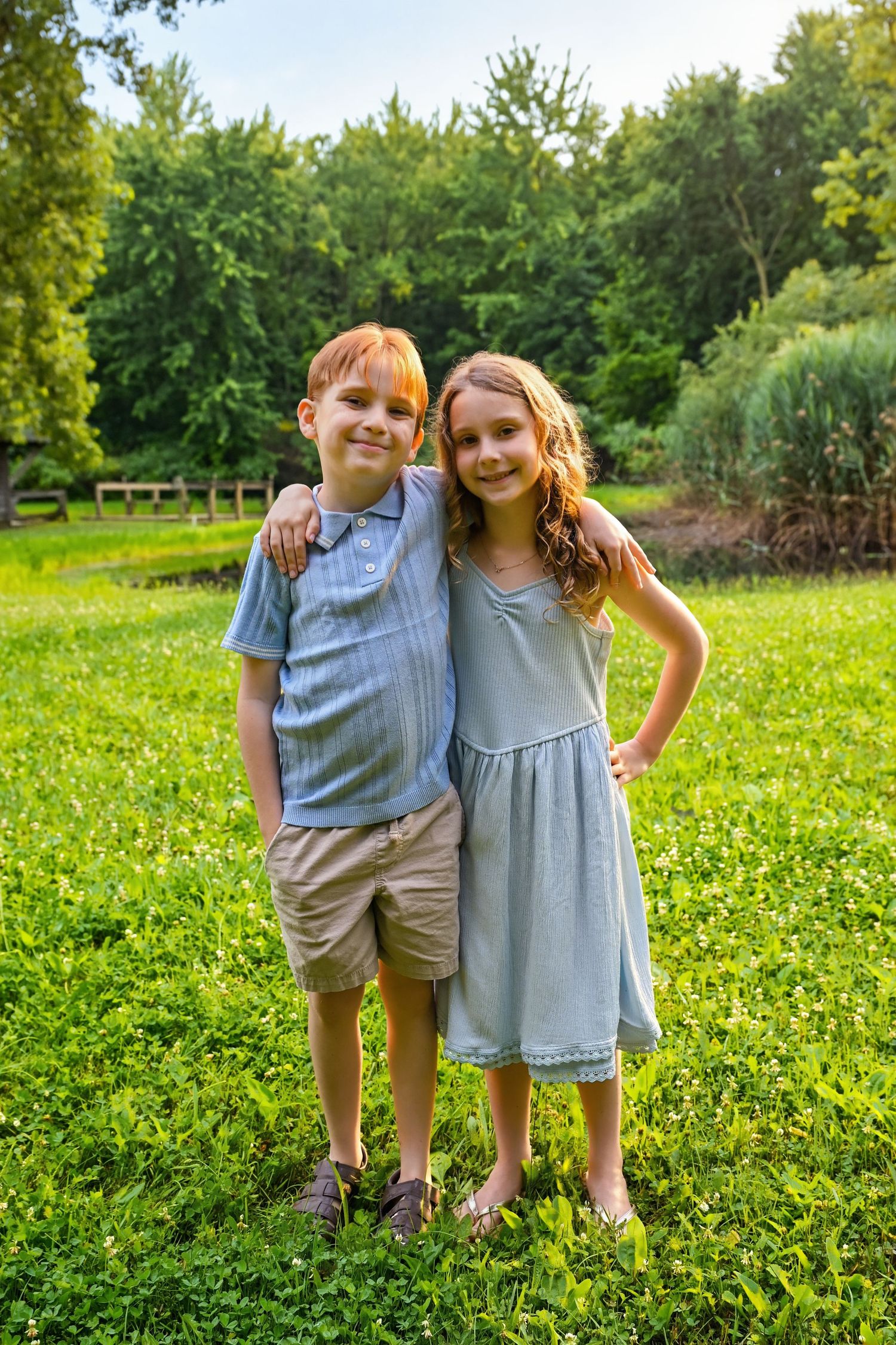 A red-haired boy and a girl in a blue dress embrace and smile together on a lush green lawn outdoors.