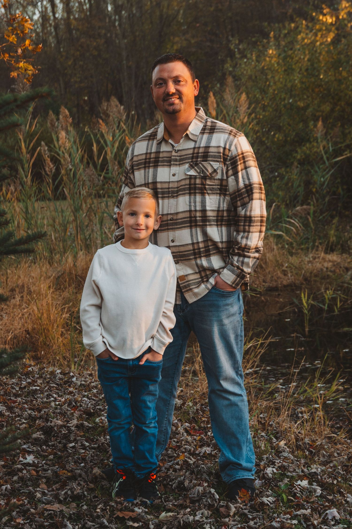A smiling father in plaid flannel shirt stands proudly with his young son outdoors in an autumn landscape.