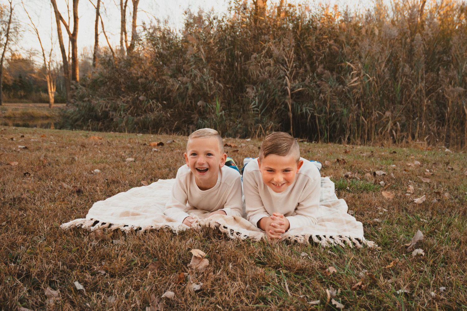 Two smiling children in cream sweatshirts lay on a white blanket in a golden autumn field laughing.
