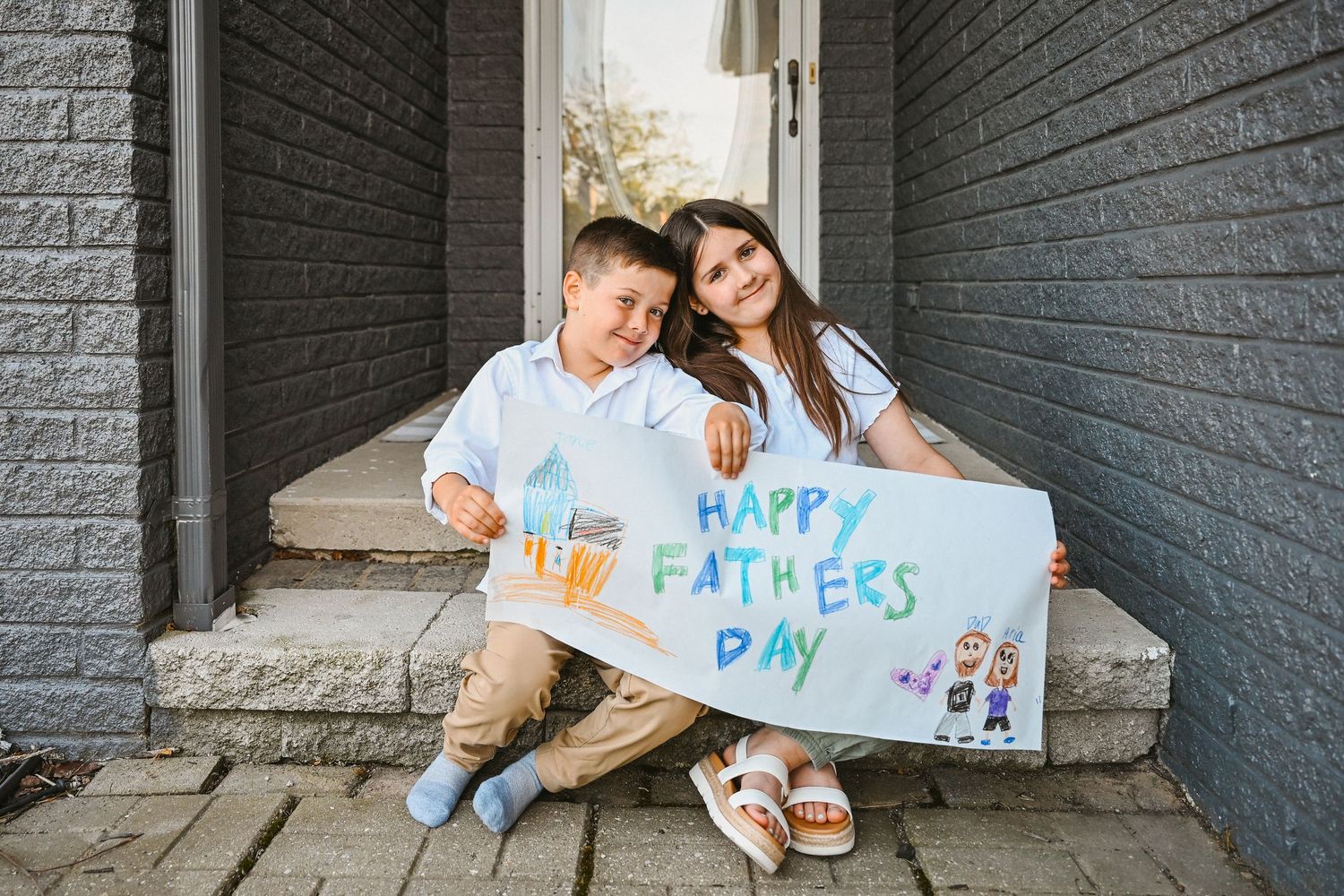 Two children sit on front porch steps proudly holding a handmade Happy Fathers Day sign.