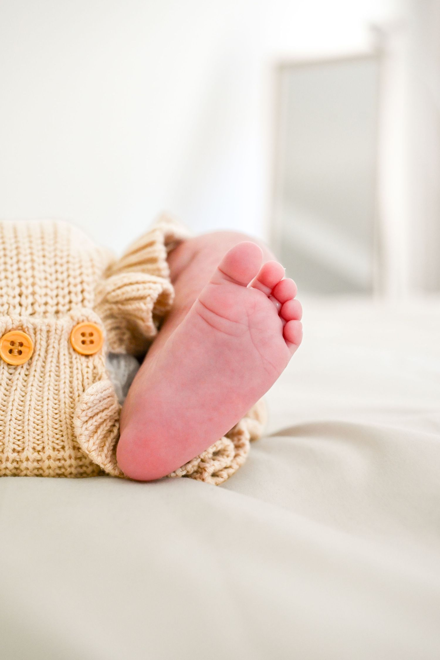 Close-up of a baby's tiny foot peeking out from a cozy beige knit sweater with orange buttons on a white bed.