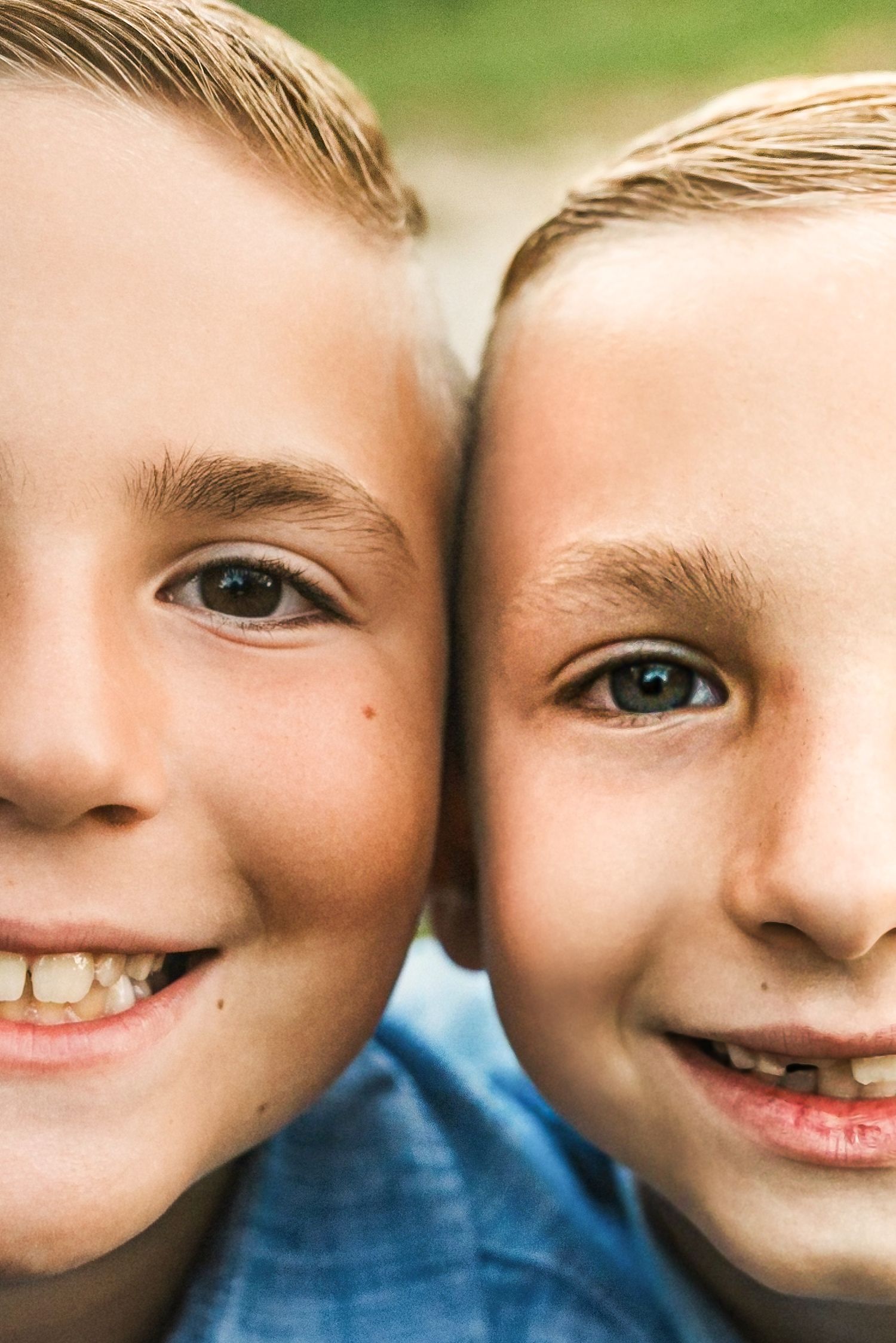 Close-up portrait of two smiling blonde boys with brown eyes posing cheek to cheek outdoors.