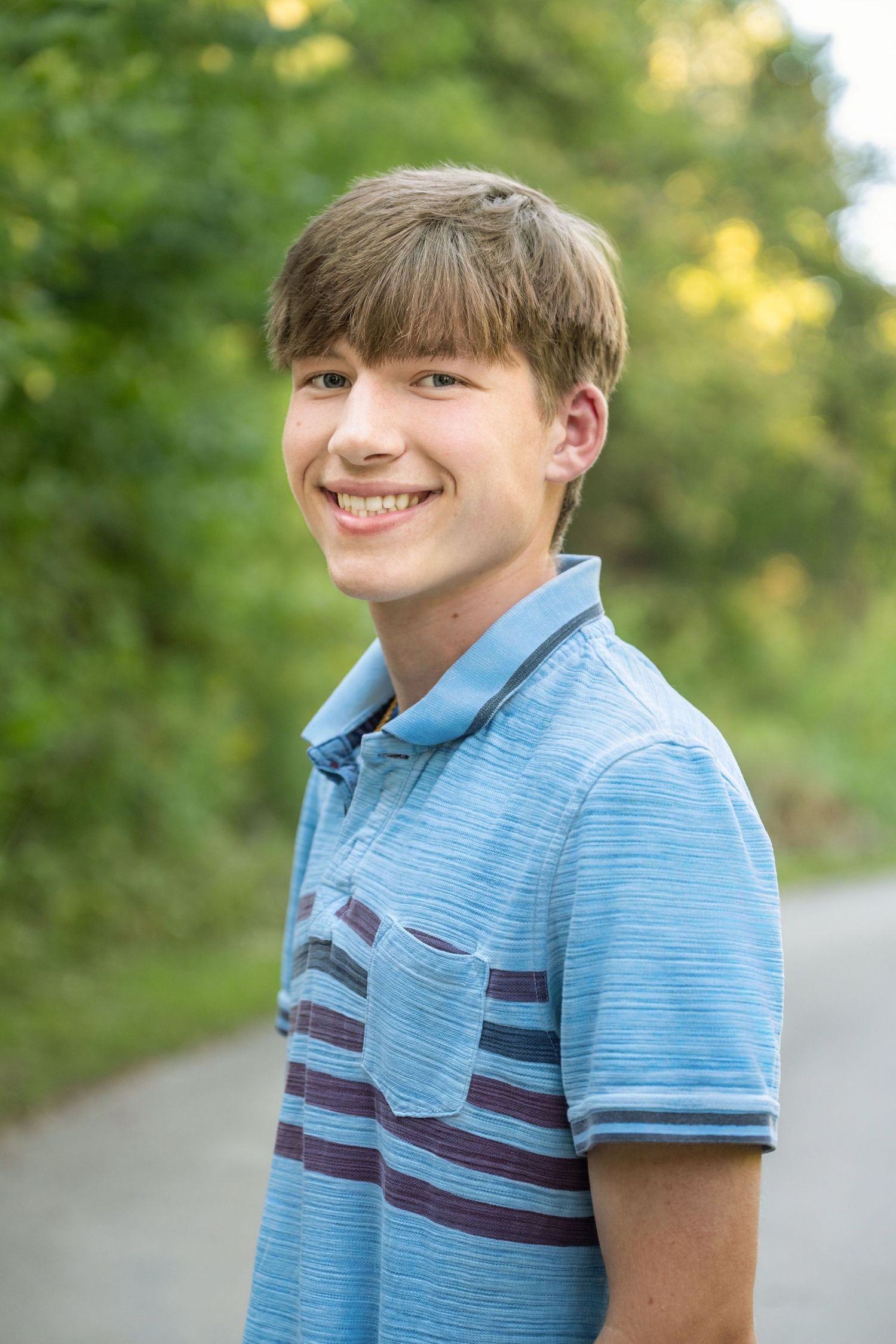 Smiling teenage boy in a blue striped polo shirt standing outdoors on a path surrounded by green trees.