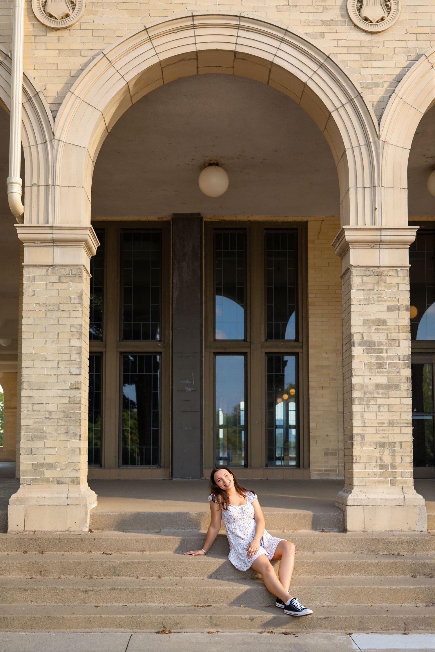 Young woman in white mini dress relaxes on steps of a grand stone building with arched entry.