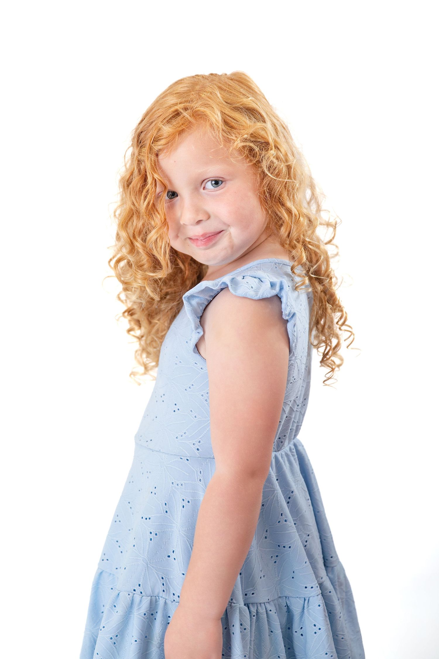 Cute little girl with long curly red hair wearing a blue dress, posing on a white background.