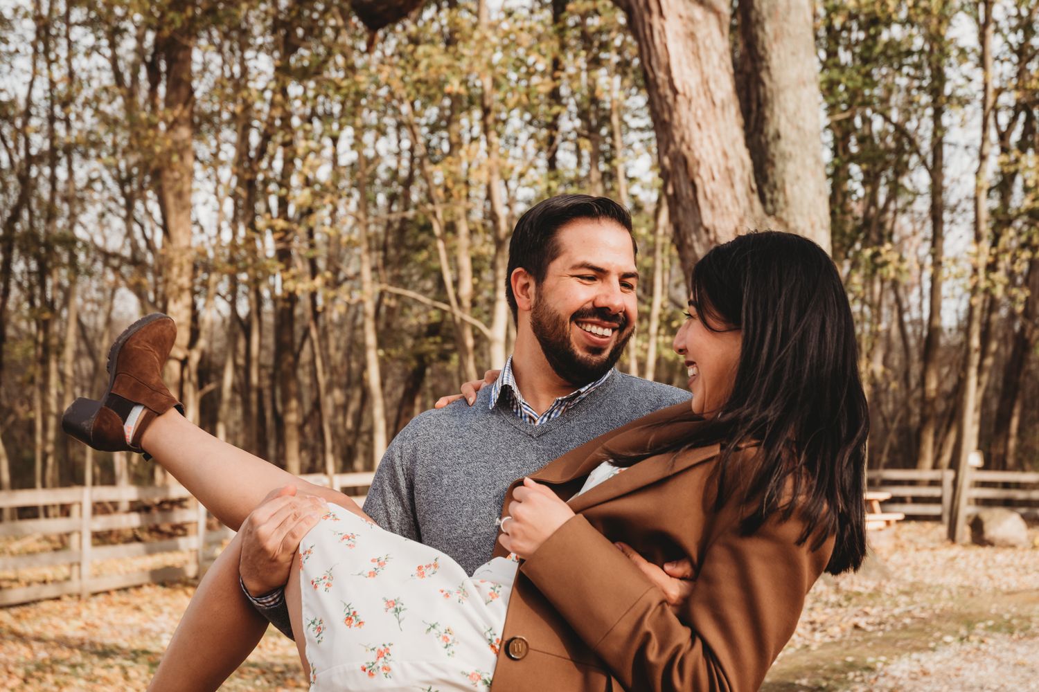 A smiling man carries a laughing woman in his arms outdoors in a sunny wooded park setting.