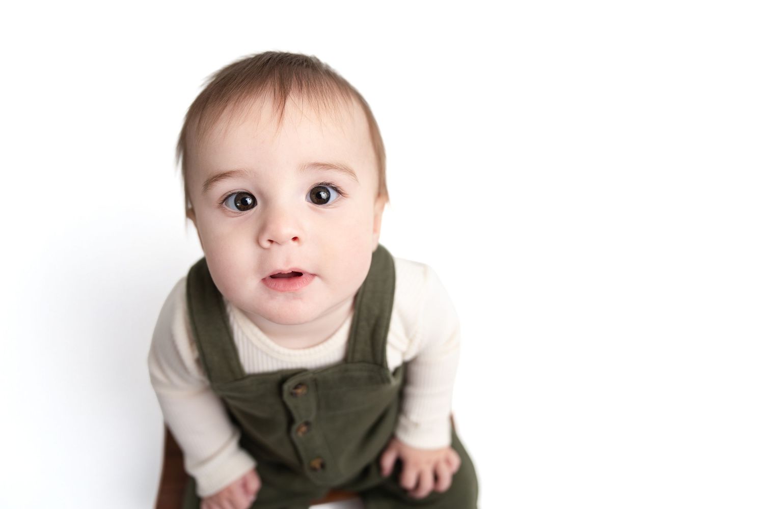Cute baby in green overalls and white shirt looks up curiously against a white background.