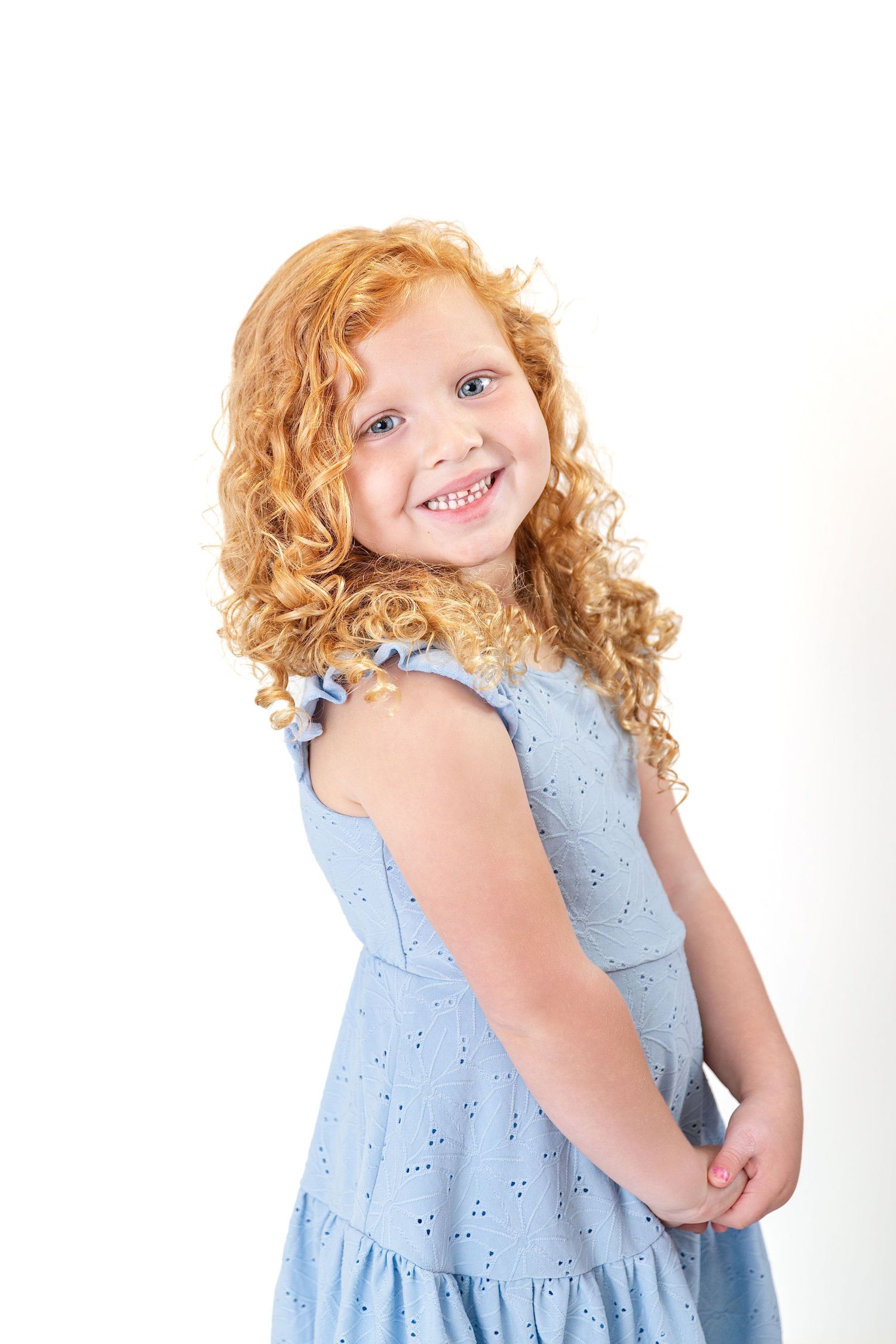Happy little girl with curly red hair in a blue eyelet dress smiling over her shoulder against white background.
