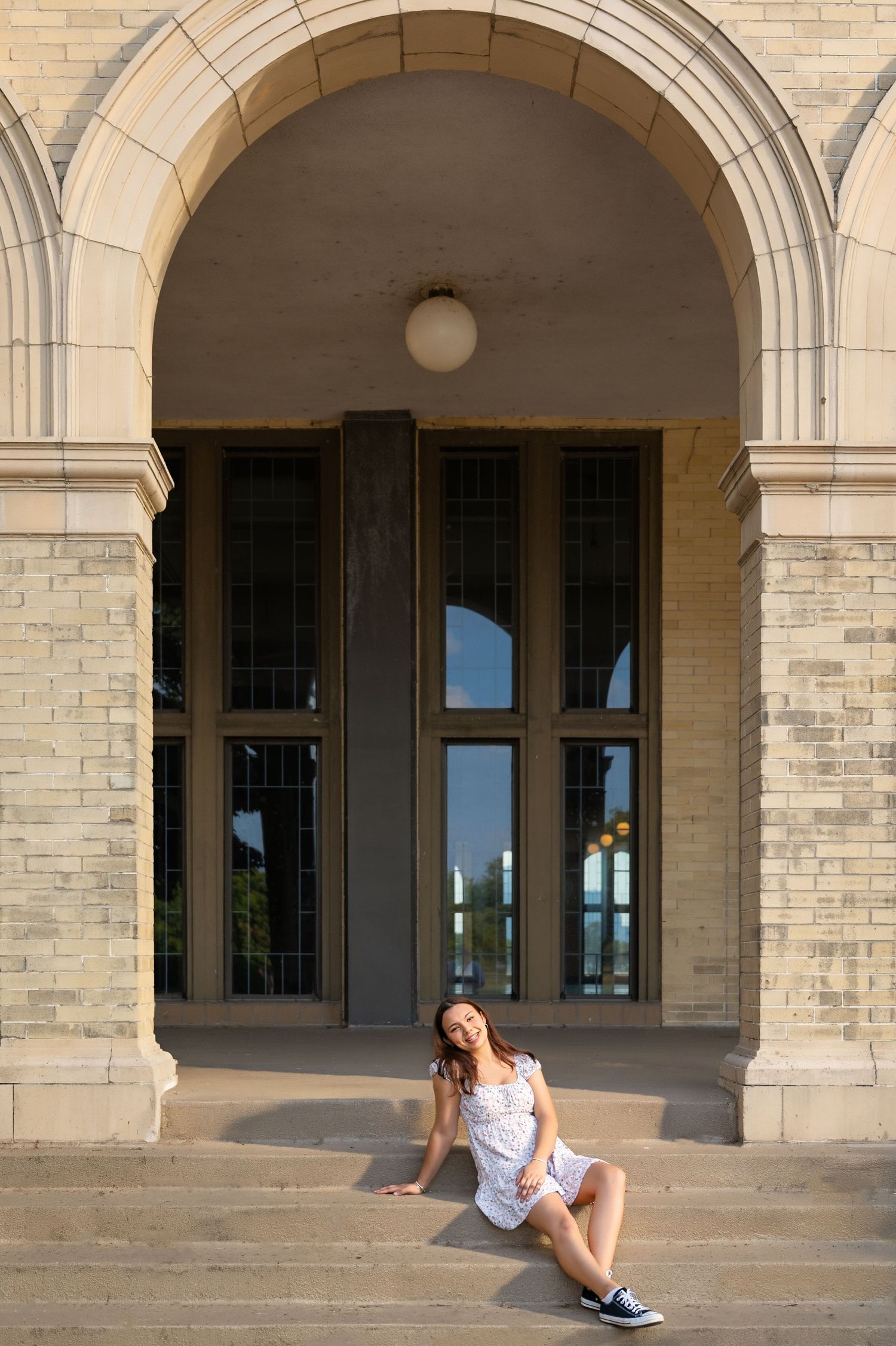 Young woman in white dress sitting on stone steps beneath an elegant arched entrance of a historic building.