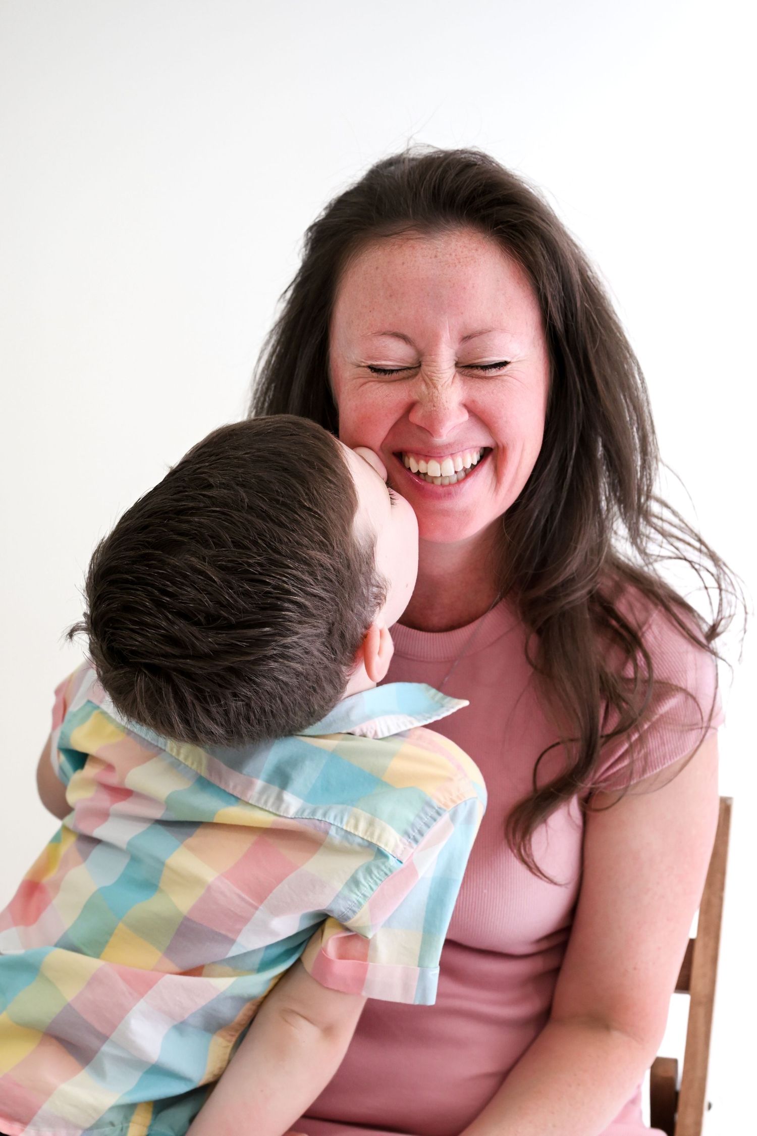A laughing mother holds her young son in a plaid shirt on her lap, sharing a joyful moment together.