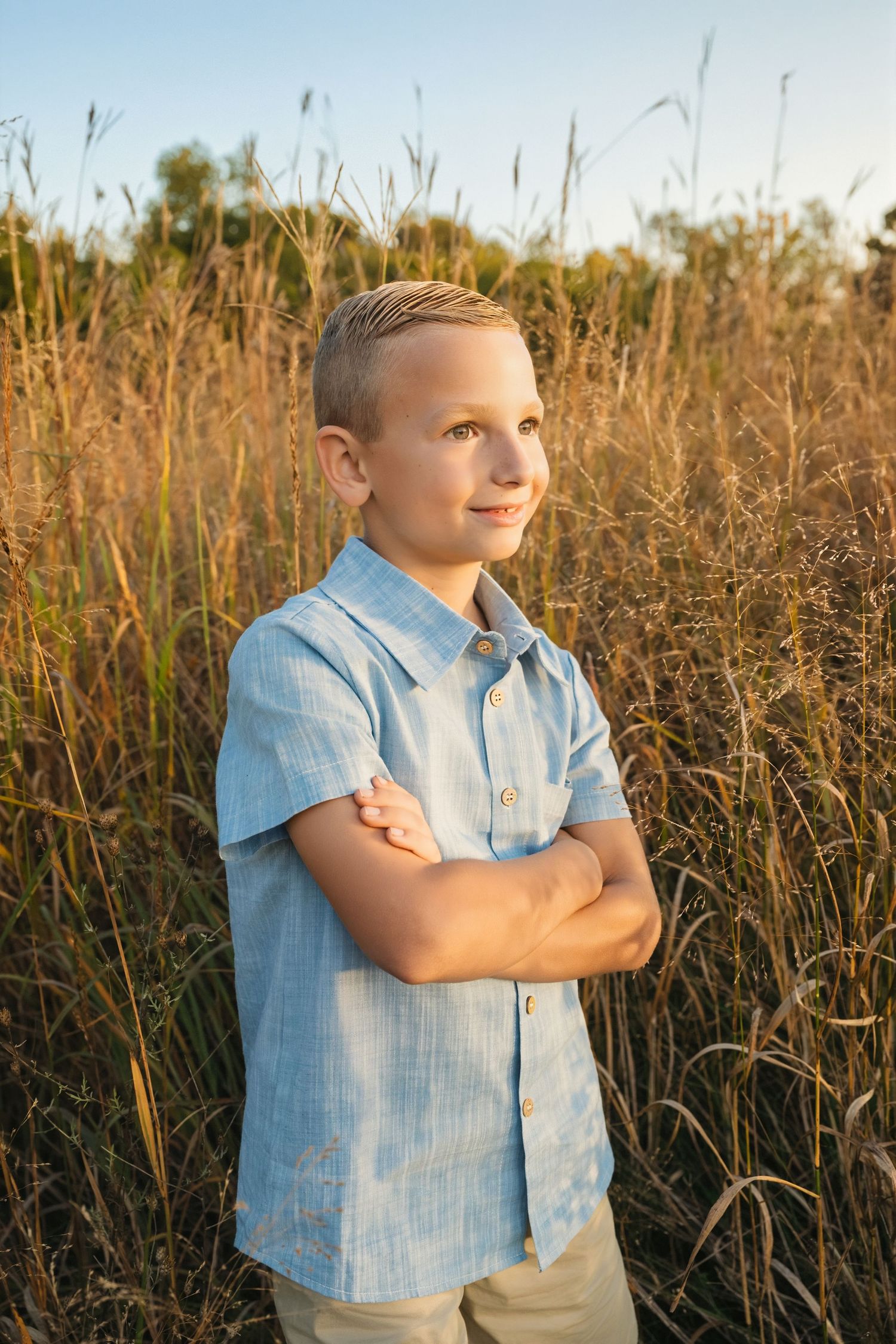 Confident young boy with crossed arms standing in golden tall grass field wearing a light blue shirt.