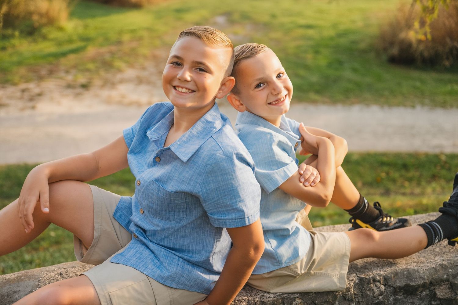 Two smiling boys in matching blue shirts sit back-to-back outdoors on a sunny day.