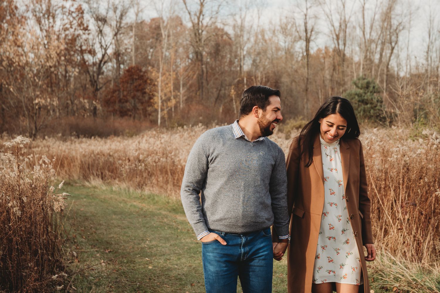 Couple holding hands and smiling while walking through an autumn field with golden grass and bare trees.