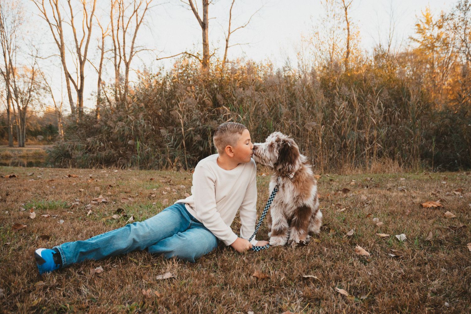 A young boy in a cream sweatshirt shares a sweet kiss with his fluffy dog outdoors in a fall setting.