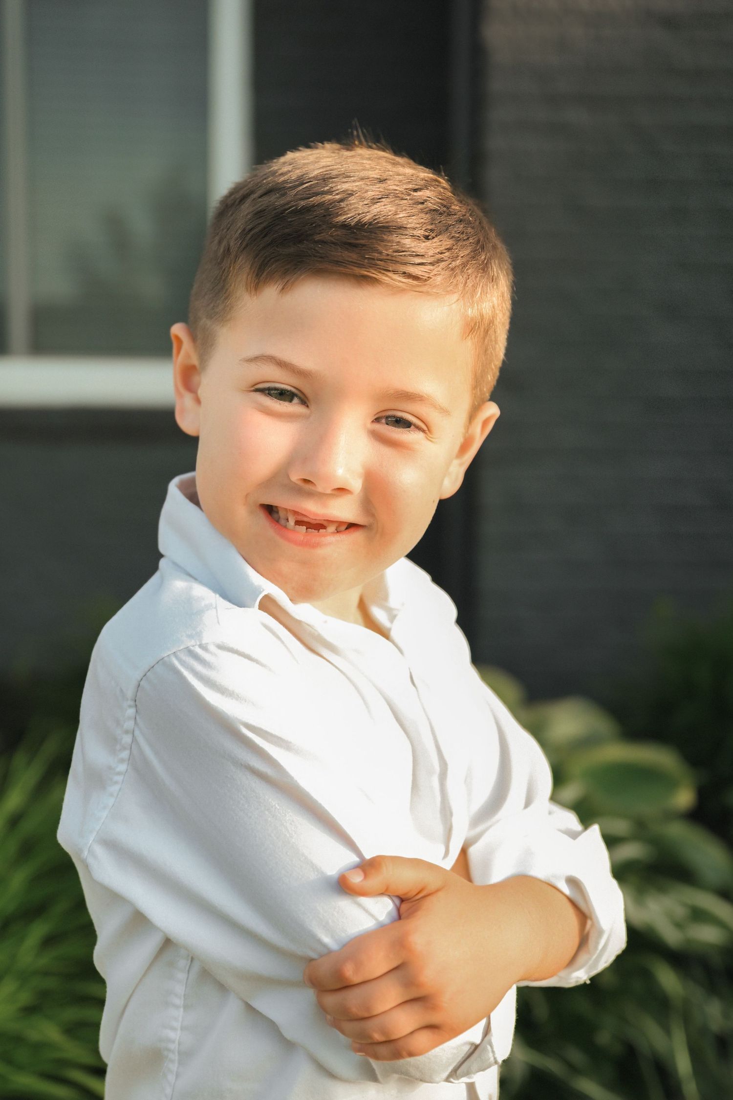 Happy young boy in white shirt stands with arms crossed smiling in a sunny backyard setting.