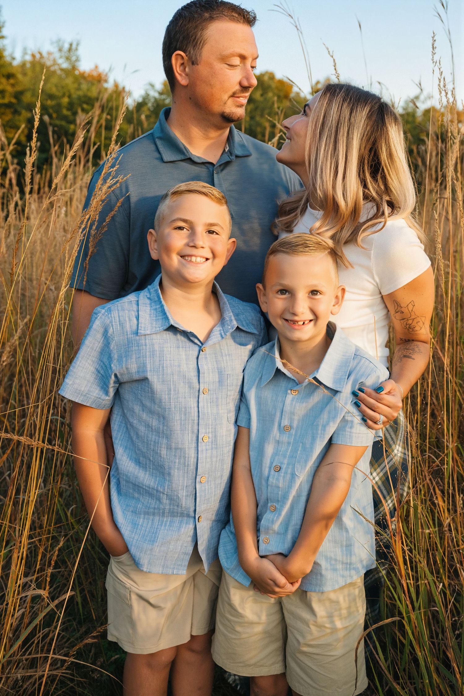 Family of four posing in golden tall grass field, two boys in matching blue shirts smiling, parents embracing behind them.